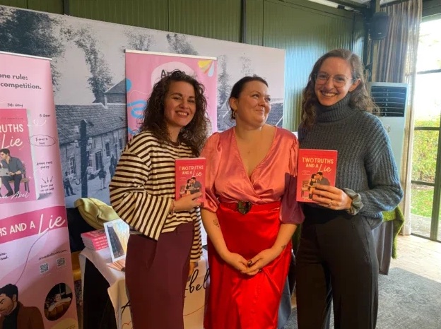 Three women smiling at a book event, two of them holding copies of a novel